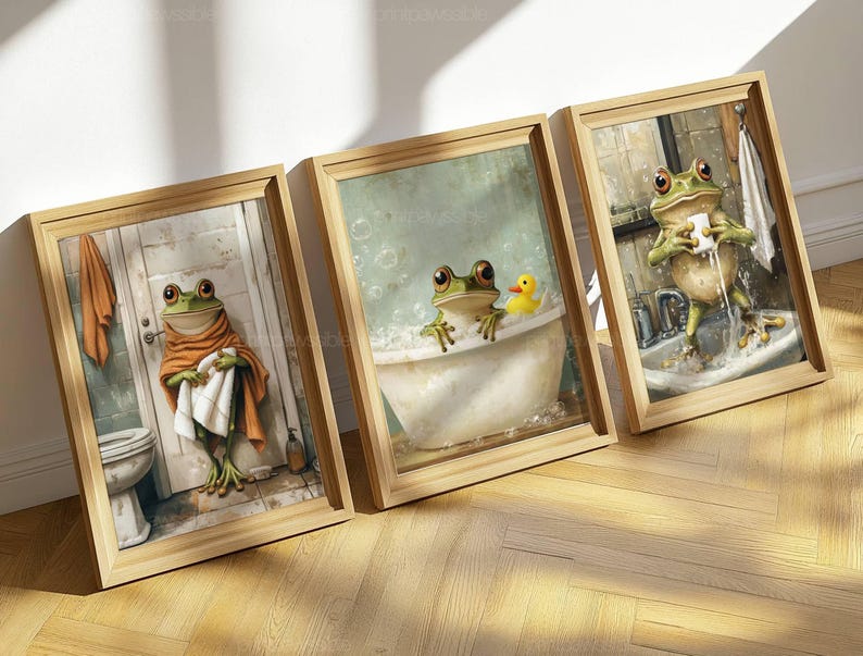 May include: Three framed art prints featuring whimsical frog illustrations in a bathroom setting. One frog is wrapped in a towel, another in a bathtub with a rubber duck, and the third washing its face at a sink. The frames are light brown.