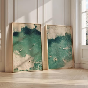 Może przedstawiać: Dwa oprawione wydruki zdjęć lotniczych surferów jadących na falach w oceanie. Fale są w żywym turkusowym kolorze, a niebo jest jasnoniebieskie. Surferzy to małe postaci w oddali.