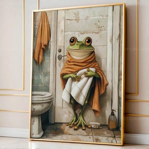 Peut inclure: Une impression d'art encadrée représente une grenouille verte dans une salle de bain. La grenouille est enveloppée dans une serviette marron et tient une serviette blanche. Des toilettes, une porte et un distributeur de savon sont également visibles. L'œuvre a un thème fantaisiste et humoristique.