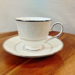 May include: A white teacup and saucer set with silver trim. The teacup features a delicate floral and dot pattern. The saucer has a matching design. The set is placed on a wooden surface.