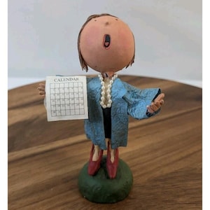 May include: A whimsical figurine of a person holding a calendar. The figure has a large, round head, wearing a blue coat and red shoes. A pearl necklace adorns the neck. The figurine stands on a green base, with the calendar visible.