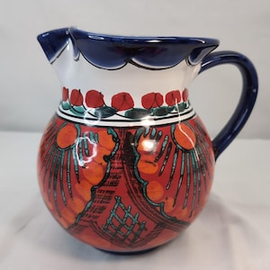 May include: A ceramic pitcher with a rounded body and a curved handle. The pitcher features a red background with orange and teal designs. The rim is white with a scalloped blue edge, and the handle is blue. Red floral accents are also present.