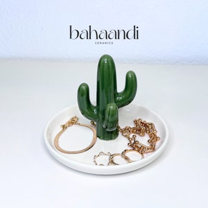 May include: A white ceramic ring dish with a green ceramic cactus sculpture in the center. The dish has gold rings and a gold chain on it. The cactus sculpture has the text "bahaandi ceramics" on it.