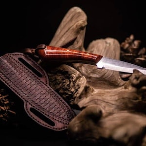 May include: A handcrafted knife with a polished wooden handle and a stainless steel blade, resting on a piece of driftwood. A brown leather sheath with a woven pattern is next to the knife. The knife is a hunting or utility knife.