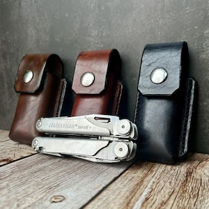 May include: A multi-tool with the text "LEATHERMAN WAVE+" is displayed with three leather sheaths in brown, dark brown, and black. The sheaths have a snap closure. The multi-tool is silver.