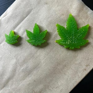 Könnte beinhalten: Drei grüne, blattförmige Gummibonbons in verschiedenen Größen liegen auf Backpapier. Die durchscheinenden Süßigkeiten weisen detaillierte Aderstrukturen und einen leicht gelblichen Farbton an den Rändern auf.