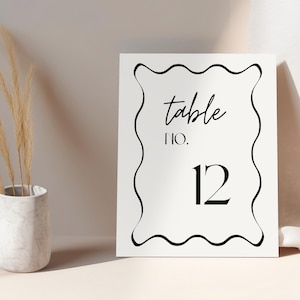 May include: A white table number card with black wavy border and text. The card reads "table NO. 12" in a stylish font. A vase with dried plants is on the left.
