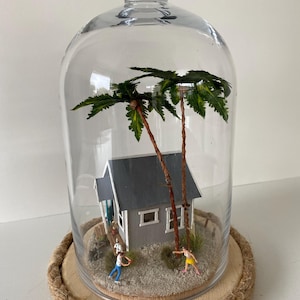 May include: A miniature beach scene with a small gray house, palm trees, and figures, all enclosed in a glass dome on a wooden base.