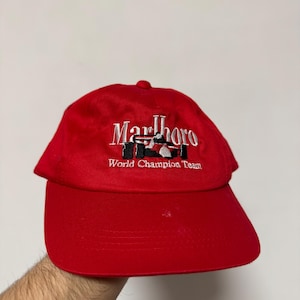 Può includere: Cappellino da baseball rosso con la scritta ricamata "Marlboro" e "World Champion Team" in bianco. Il cappellino presenta una piccola immagine di un'auto da corsa.