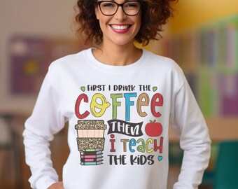 May include: White sweatshirt with a colourful graphic that says "First I drink the coffee then I teach the kids". The graphic includes a coffee cup with a leopard print design, a stack of books, and an apple.