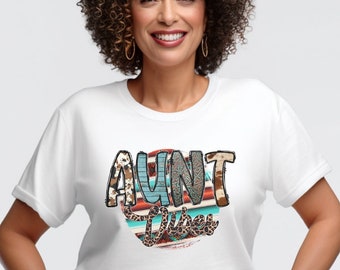 May include: White t-shirt featuring a colourful graphic design that reads "Aunt Vibes". The graphic incorporates a turquoise, red, and brown colour scheme with a leopard print and paisley pattern.
