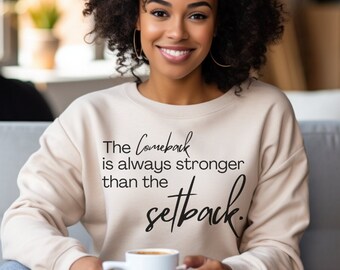 May include: A beige sweatshirt with the text "The Comeback is always stronger than the setback" in black script. A person is wearing the sweatshirt and holding a white mug.