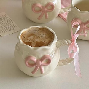 May include: Cream-colored ceramic mugs with pink bow accents. The mugs have a unique shape and twisted handles. One mug is filled with coffee. The mugs are arranged on a white surface, suggesting a cozy setting.