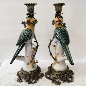 May include: A pair of ceramic candle holders featuring colorful parrots perched on white tree stumps. Each piece has a brass-toned metal base and a matching brass-toned candle cup at the top. The parrots have yellow heads, green wings, and long tails.