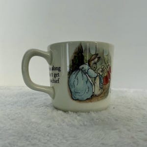 May include: A white ceramic mug with a curved handle. The mug features a colorful illustration of Peter Rabbit and other characters from the children's book. Text is printed on the side of the mug.