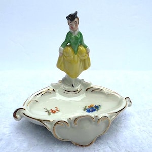 May include: A vintage porcelain trinket dish featuring a figurine of a woman in a green and yellow dress. The dish is white with gold trim and floral accents. The figurine is wearing a black hat.