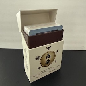 May include: A cream-colored card game box with an open lid, revealing a stack of cards inside. The box features a central gold seal with a lantern icon, surrounded by small animal silhouettes. Text on the box reads: Timeless Tales, Lasting Lessons, Family Fun, Ages 6+, 2-4 Players.