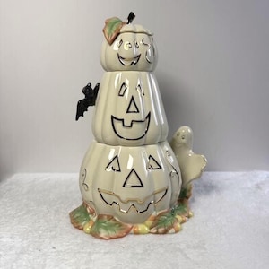 May include: A ceramic stack of three cream-colored pumpkins with gold-trimmed jack-o'-lantern faces. A small black bat is attached to the side of the middle pumpkin, and a white ghost figure stands at the base, which is decorated with painted leaves.
