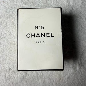 May include: A rectangular Chanel No 5 perfume box with a cream-colored front panel and black border. The text N&deg; 5, CHANEL, and PARIS is printed in black on the front. The box has a clean, minimalist design with sharp edges and a vertical orientation.