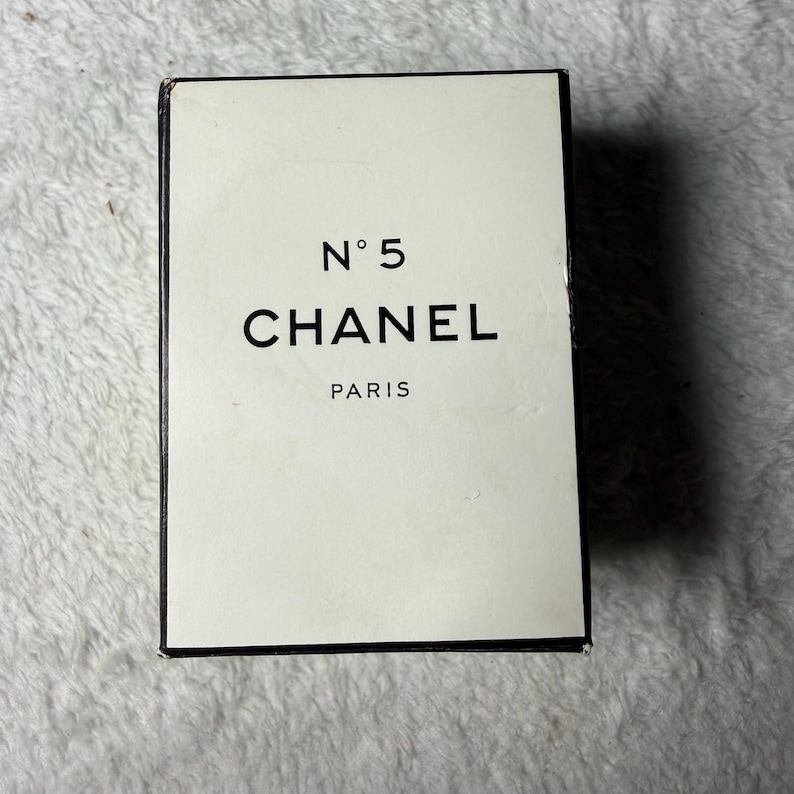 May include: A rectangular Chanel No 5 perfume box with a cream-colored front panel and black border. The text N&deg; 5, CHANEL, and PARIS is printed in black on the front. The box has a clean, minimalist design with sharp edges and a vertical orientation.
