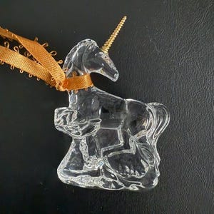 May include: A clear glass unicorn ornament with a gold horn. The ornament is hanging from a brown ribbon.