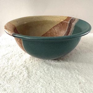 May include: A round ceramic bowl featuring a multi-colored glaze design. The exterior and interior rim display a deep teal color, while the inner basin transitions into bands of warm beige and textured brown, creating a distinct color-blocked appearance.