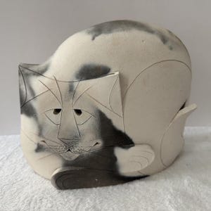 May include: A ceramic sculpture of a curled cat with a matte off-white finish and dark gray smoke-like markings. The cat features etched geometric lines defining its face, ears, and paws, with a smooth, rounded body shape and a small tail.