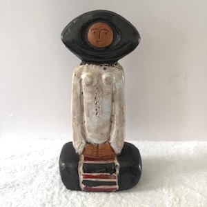 May include: A ceramic figurine featuring a central white torso with a textured surface, topped by a large black oval headpiece with a small brown circular face. The base is black with horizontal red and white stripes at the center front.