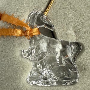 May include: A clear glass unicorn figurine with a gold horn. The unicorn is standing on a base and is tied with a gold ribbon.