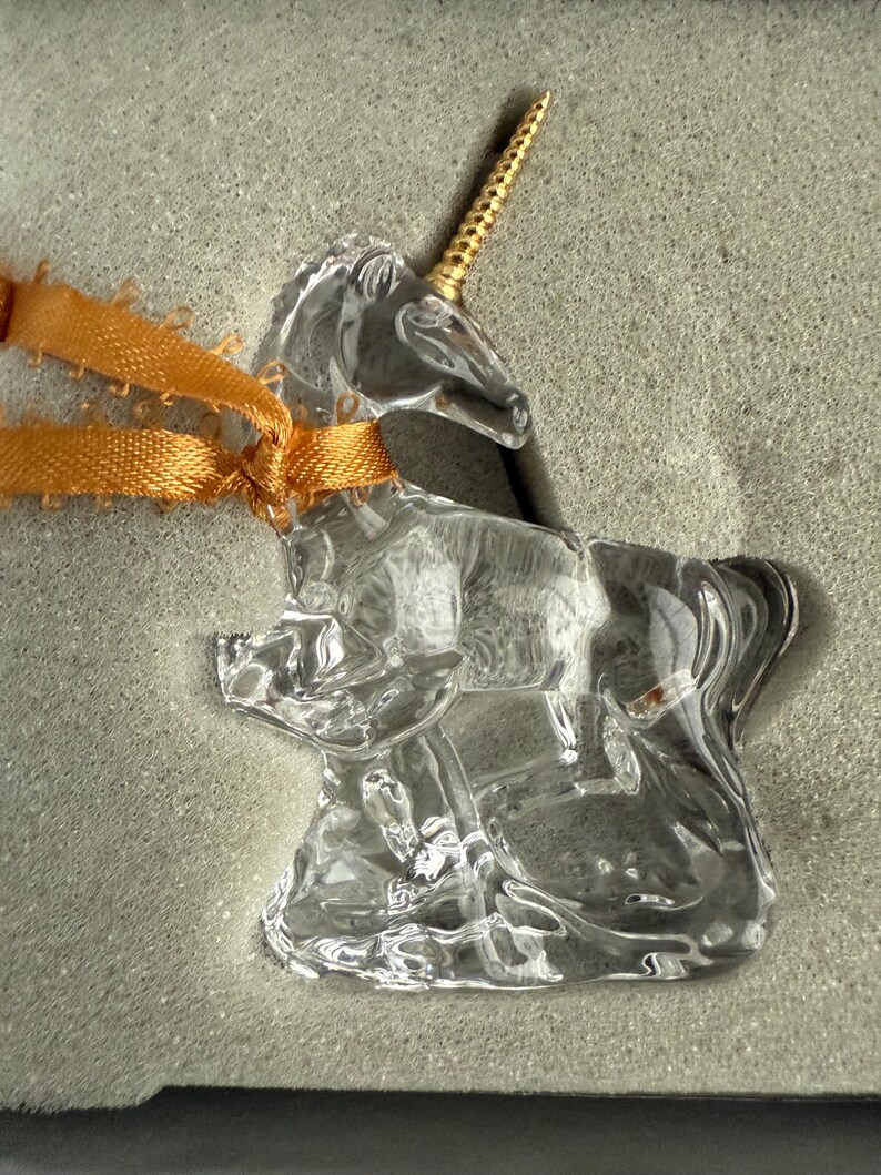 May include: A clear glass unicorn figurine with a gold horn. The unicorn is standing on a base and is tied with a gold ribbon.