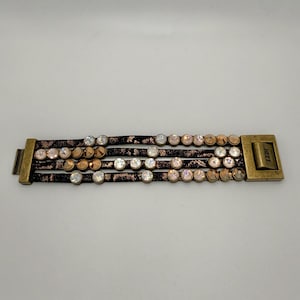May include: A multi-strand bracelet featuring four dark brown textured leather bands adorned with round, faceted clear and amber-toned crystals. The bracelet is secured by rectangular antique brass-toned metal clasps, with the word HEET engraved.