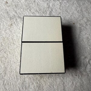 May include: A rectangular, hinged compact case featuring a matte, off-white finish on the top and bottom panels. The exterior edges are framed in a contrasting black border, creating a clean, geometric design with a central horizontal seam.