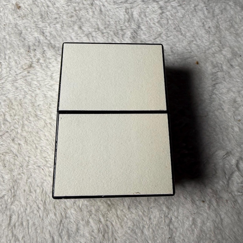 May include: A rectangular, hinged compact case featuring a matte, off-white finish on the top and bottom panels. The exterior edges are framed in a contrasting black border, creating a clean, geometric design with a central horizontal seam.