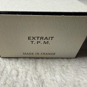 May include: A rectangular off-white cardboard box with black text printed on the front. The text reads EXTRAIT T. P. M. followed by the words MADE IN FRANCE. The box has a smooth, matte finish and a clean, simple design structure.
