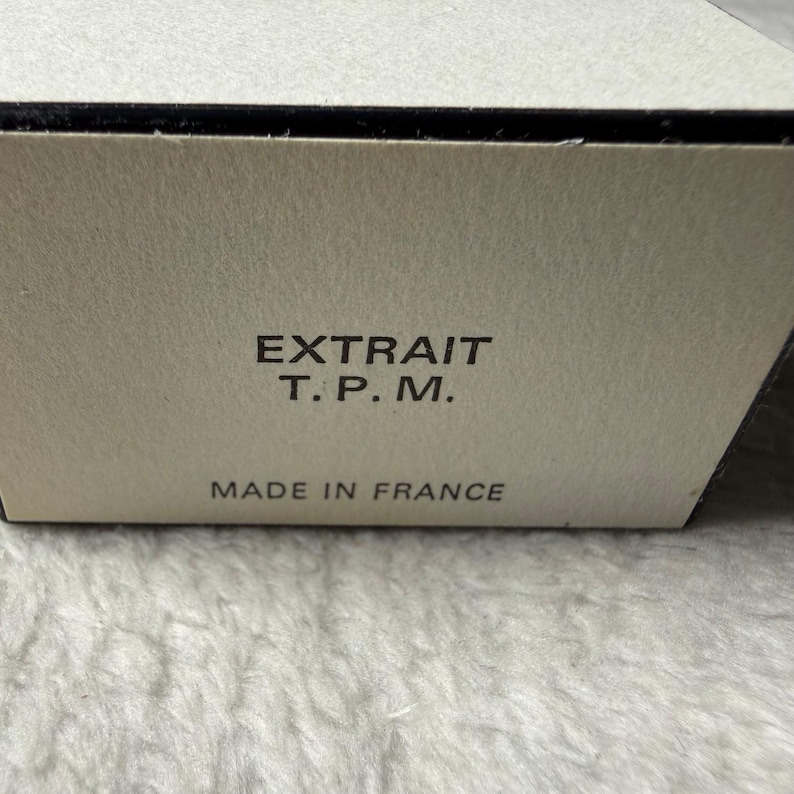 May include: A rectangular off-white cardboard box with black text printed on the front. The text reads EXTRAIT T. P. M. followed by the words MADE IN FRANCE. The box has a smooth, matte finish and a clean, simple design structure.