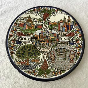 May include: A round decorative ceramic plate featuring a colorful illustration of the Holy Land. The design includes labeled sections for Nazareth, Bethlehem, and Jerusalem, surrounded by floral patterns, grape clusters, and mosaic-style art.