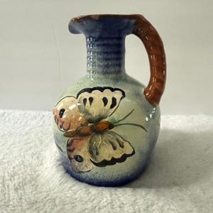 May include: A small, hand-painted ceramic pitcher with a butterfly design. The pitcher features a blue rim and handle, with a light green body. The butterfly has brown, tan, and black details. The handle is brown.