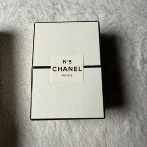 May include: A rectangular off-white box with a black border and a central label featuring the text N&deg;5 CHANEL PARIS. The box has a horizontal black line running across the middle, separating the top and bottom sections of the textured packaging.