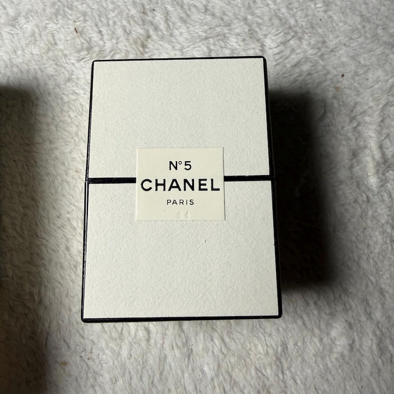 May include: A rectangular off-white box with a black border and a central label featuring the text N&deg;5 CHANEL PARIS. The box has a horizontal black line running across the middle, separating the top and bottom sections of the textured packaging.