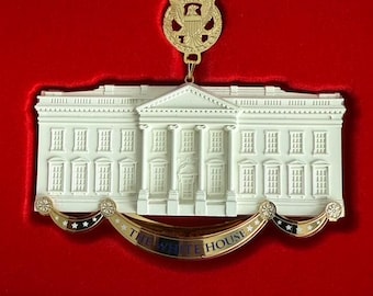 Puede incluir: Adorno de cerámica blanca de la Casa Blanca con detalles dorados y una pancarta que dice "The White House". El adorno está colgado de una cinta dorada con un emblema de águila dorada.