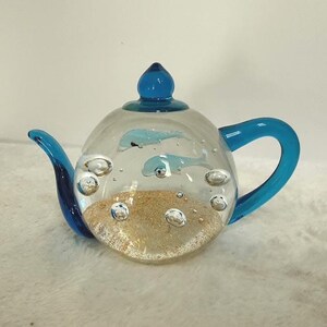 May include: A clear glass teapot with blue accents, featuring a sand-like base and two blue dolphin figures inside. The teapot has a blue handle, spout, and lid, creating a whimsical ocean-themed decorative piece.