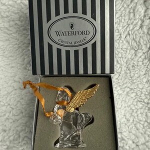 Może przedstawiać: Choinka z kryształu Waterford Crystal Jewels w pudełku. Choinka jest wykonana z kryształu i ma złote skrzydła. Pudełko jest czarne w białe paski i zawiera logo Waterford Crystal Jewels.