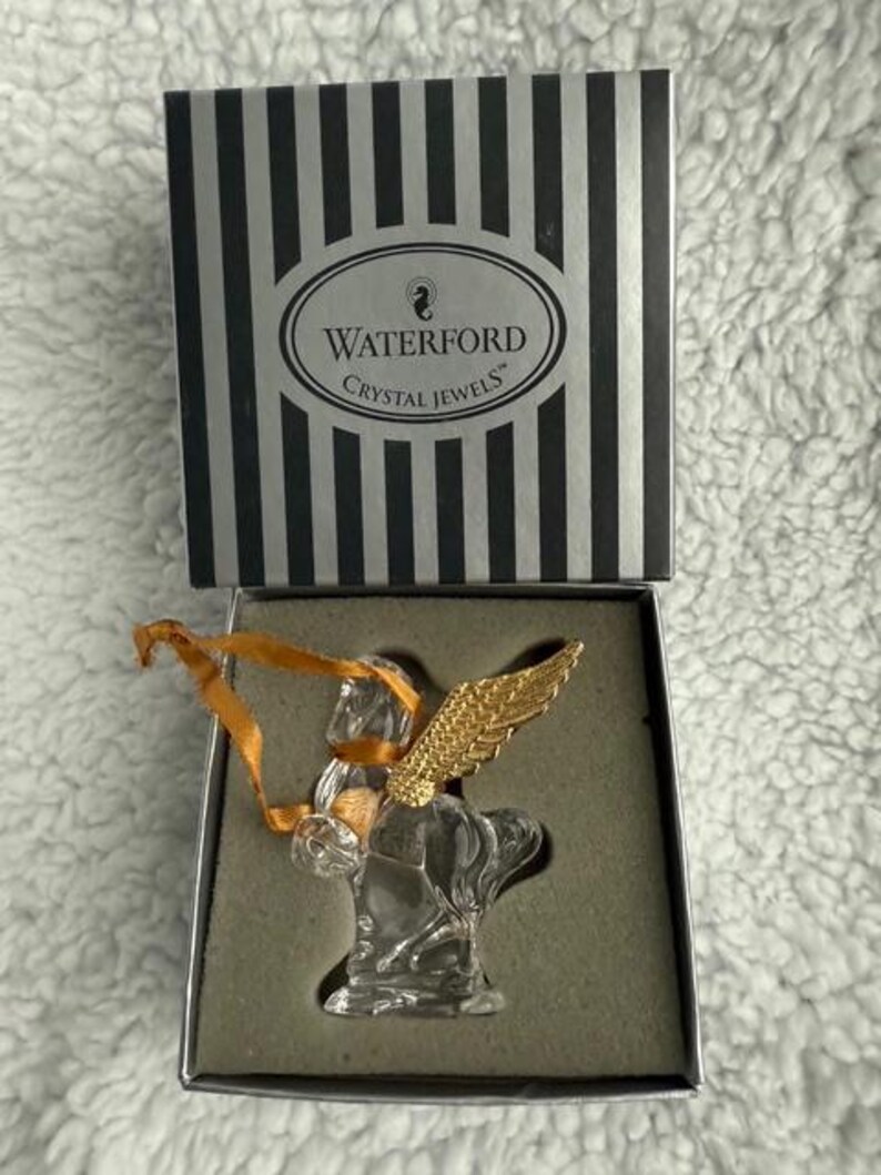 Może przedstawiać: Choinka z kryształu Waterford Crystal Jewels w pudełku. Choinka jest wykonana z kryształu i ma złote skrzydła. Pudełko jest czarne w białe paski i zawiera logo Waterford Crystal Jewels.