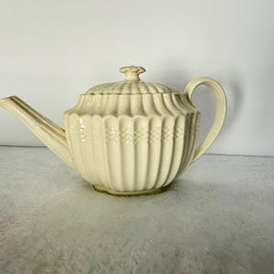 May include: Cream-colored ceramic teapot with a lid and handle. The teapot has a ribbed design with a decorative band around the middle. The spout is long and curved. The lid is topped with a small, flower-shaped knob.
