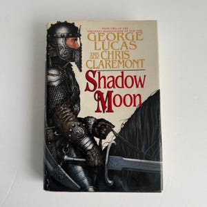 May include: A book cover featuring a knight in armor riding a black horse. The title of the book is "Shadow Moon" by George Lucas and Chris Claremont.