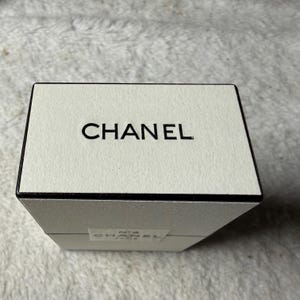 May include: A rectangular off-white box with a black border and the word CHANEL printed in black on the top surface. A smaller white label on the side of the box features the text N&deg;5 CHANEL PARIS in black lettering against a white background.