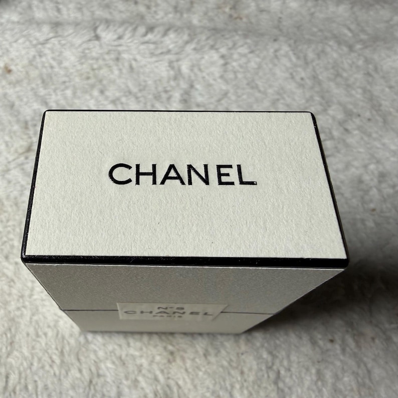 May include: A rectangular off-white box with a black border and the word CHANEL printed in black on the top surface. A smaller white label on the side of the box features the text N&deg;5 CHANEL PARIS in black lettering against a white background.