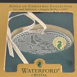 May include: A Waterford Crystal ornament from the Songs of Christmas Collection, Second Edition - Jingle Bells - 1997. The ornament is a silver oval with a depiction of a person riding in a sleigh pulled by reindeer. The ornament is in a green and gold box with the Waterford Crystal logo.