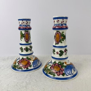 May include: Pair of ceramic candle holders with a white base, adorned with colorful floral and leaf designs. The candle holders feature a blue rim and accents of pink, orange, and green. The design is repeated on both candle holders.