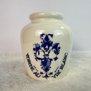 May include: A white ceramic jar with a blue floral design and the words "Forteau Vin Blanc" in blue. The jar has a rounded body and a small, slightly flared rim. The text includes "5% oz" and "160g".
